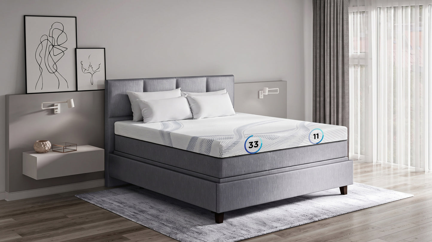 A gray upholstered bed frame with a white mattress, shown in a modern bedroom setting with a gray headboard, white pillows, wall-mounted bedside tables, framed wall art, and a window with sheer curtains.